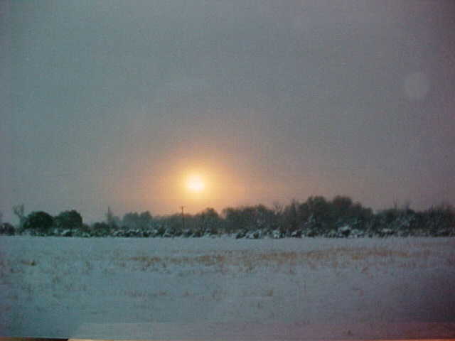 Misty winter sunset with the sun visible in the clouds and snow over the open field.